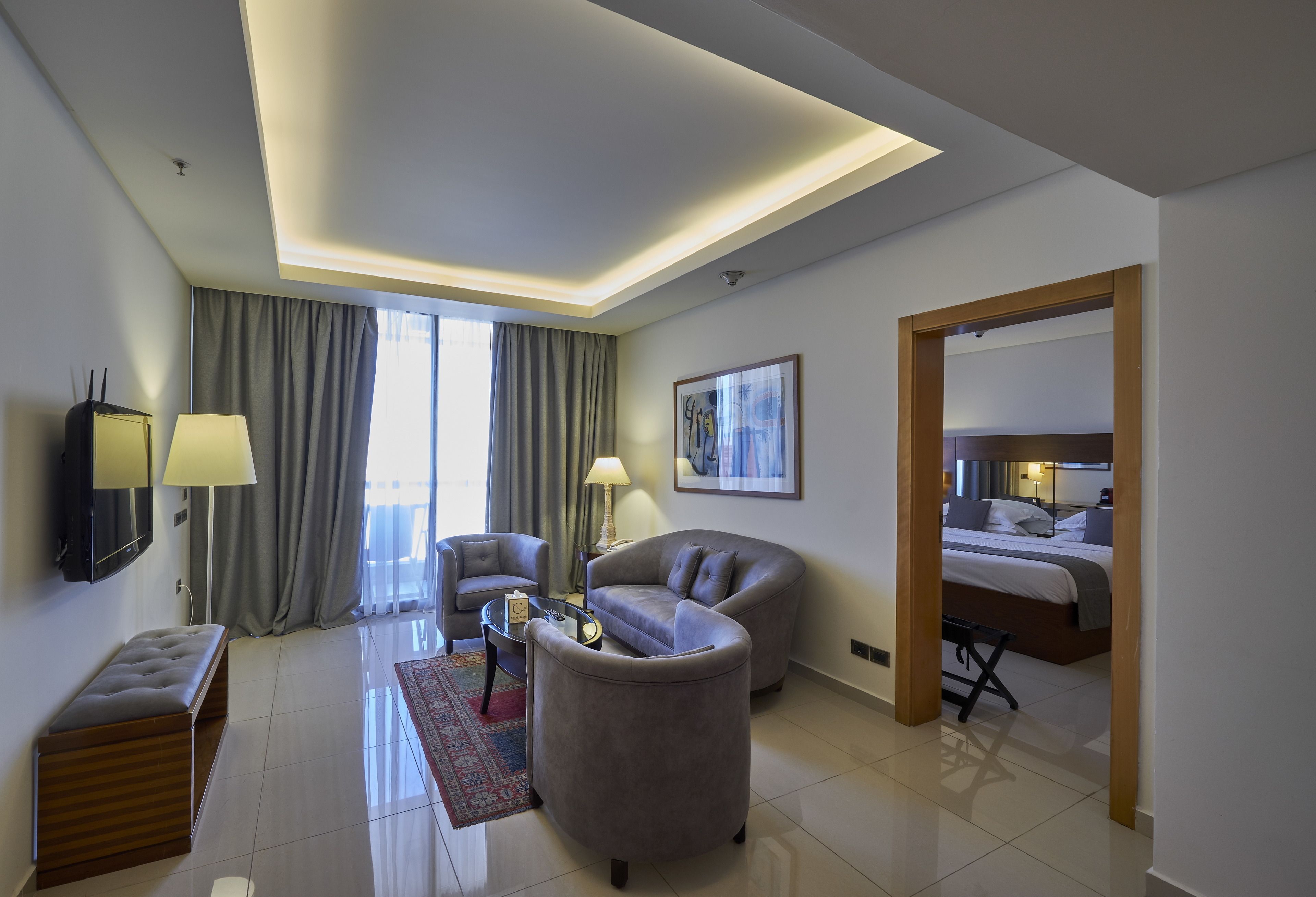 A modern hotel room with a sofa, armchair, and a small table. There is a bed with a wooden headboard and a large mirror. The room has a ceiling with recessed lighting and a view of the outdoors through large windows with curtains.