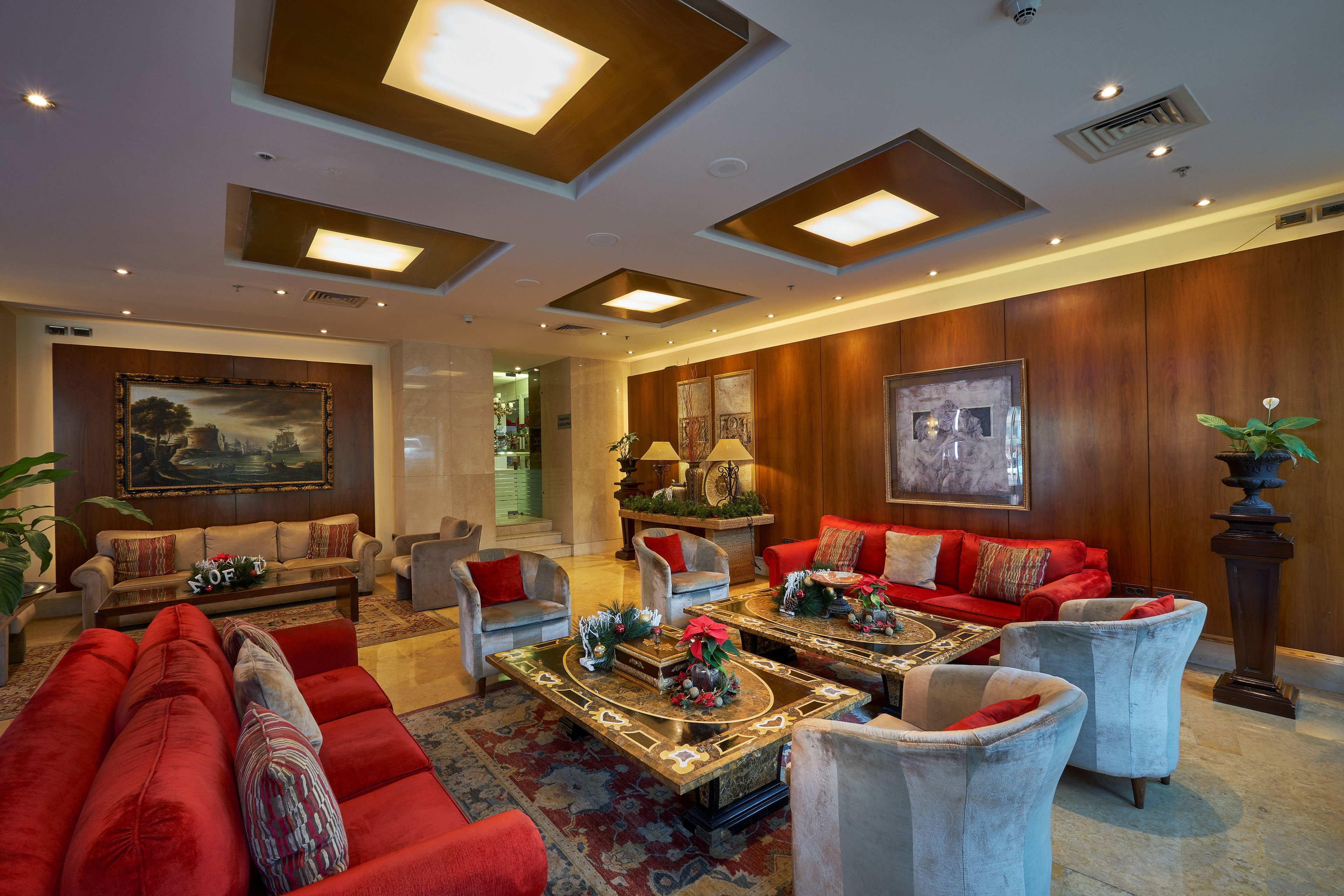 Un amplio salón con un sofá rojo grande, varios sillones y una mesa de centro. Hay luces de techo y un cuadro grande en la pared.