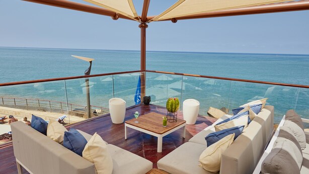 Eine Terrasse mit Holztisch, weißen Sofas und einem Glasgeländer mit Blick aufs Meer.