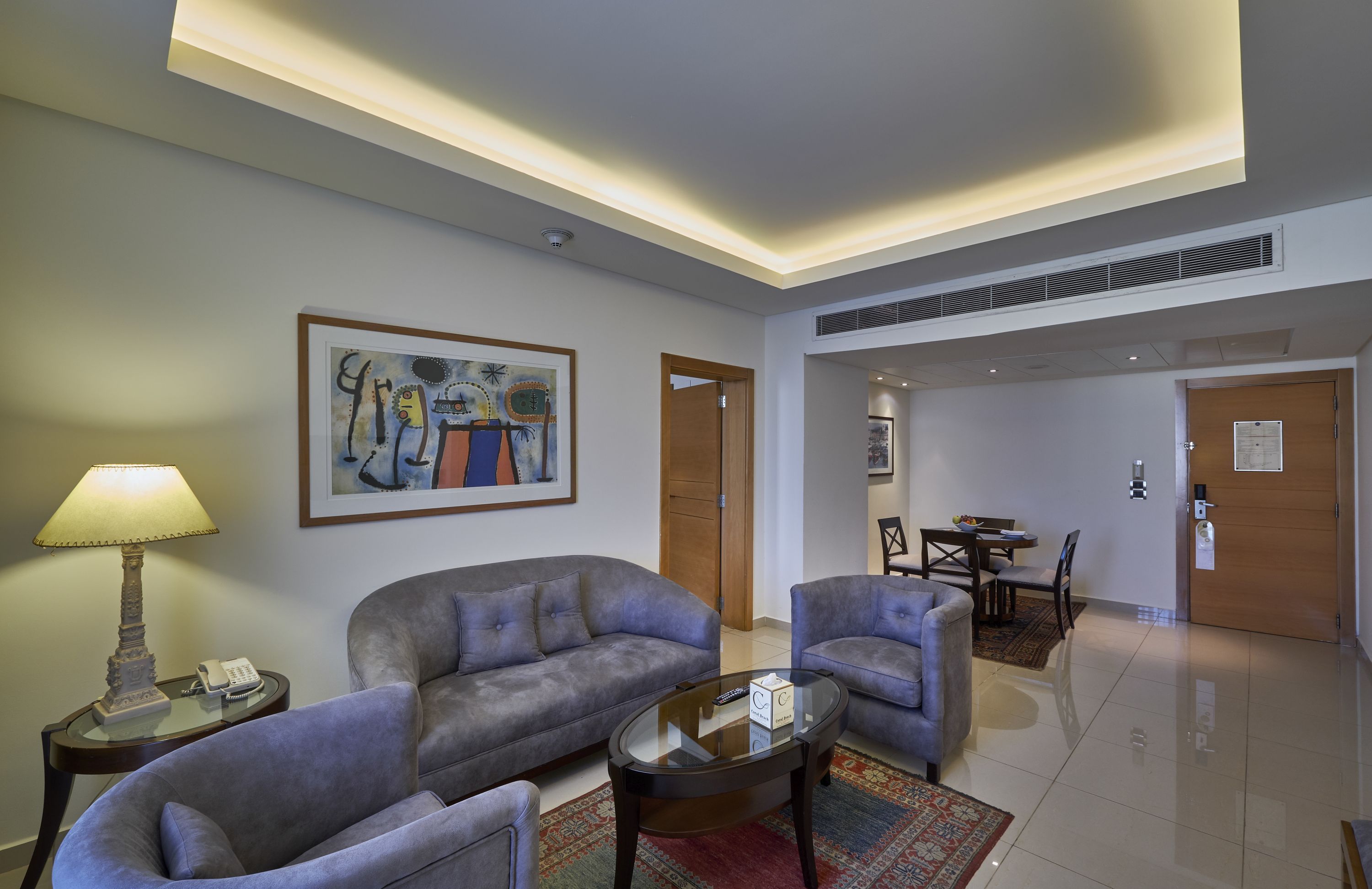 Una habitación de hotel moderna con un sofá gris, sillones, una mesa de centro de vidrio y un cuadro en la pared.