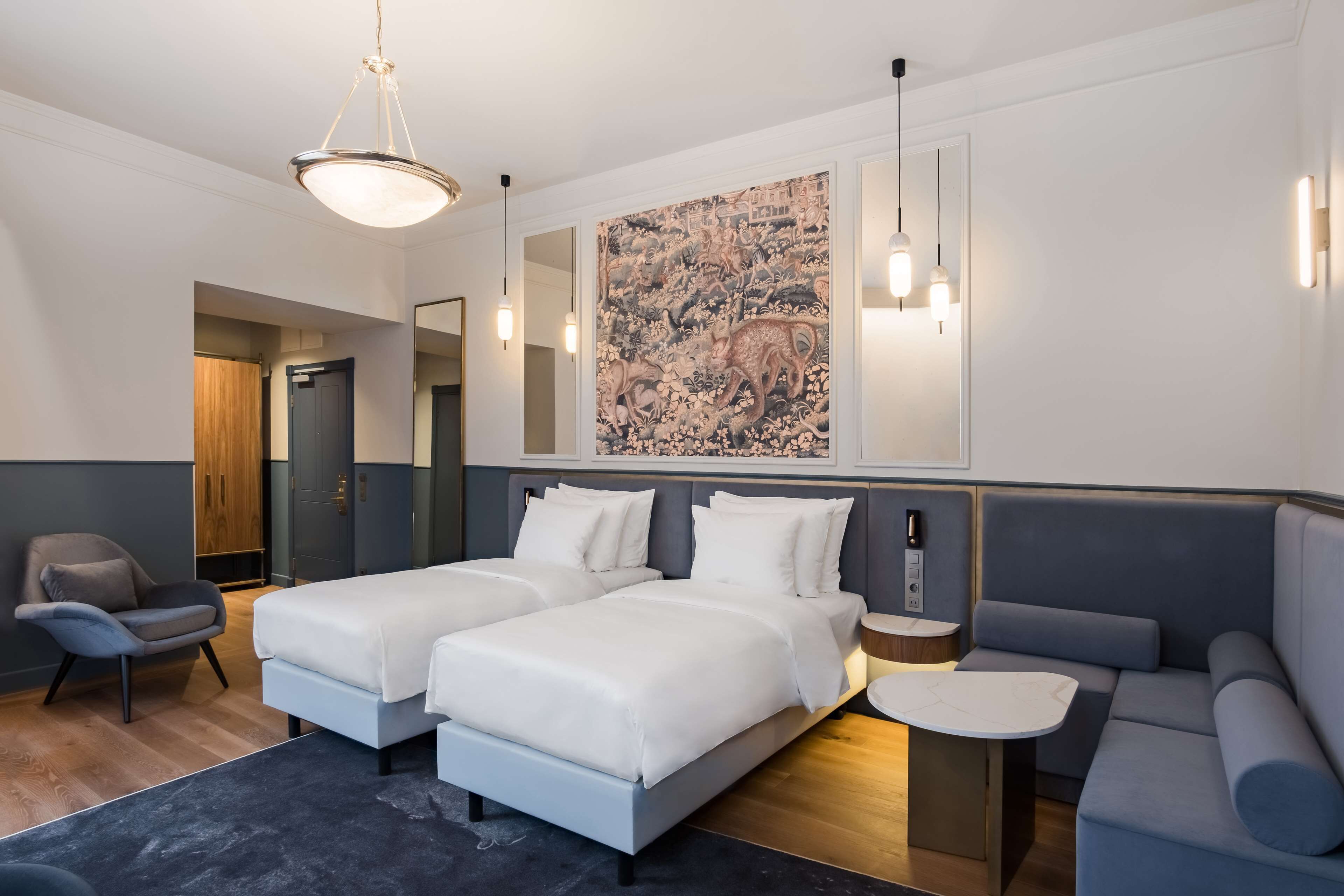 A modern hotel room with two beds, a sofa, a small table, and a large mirror.