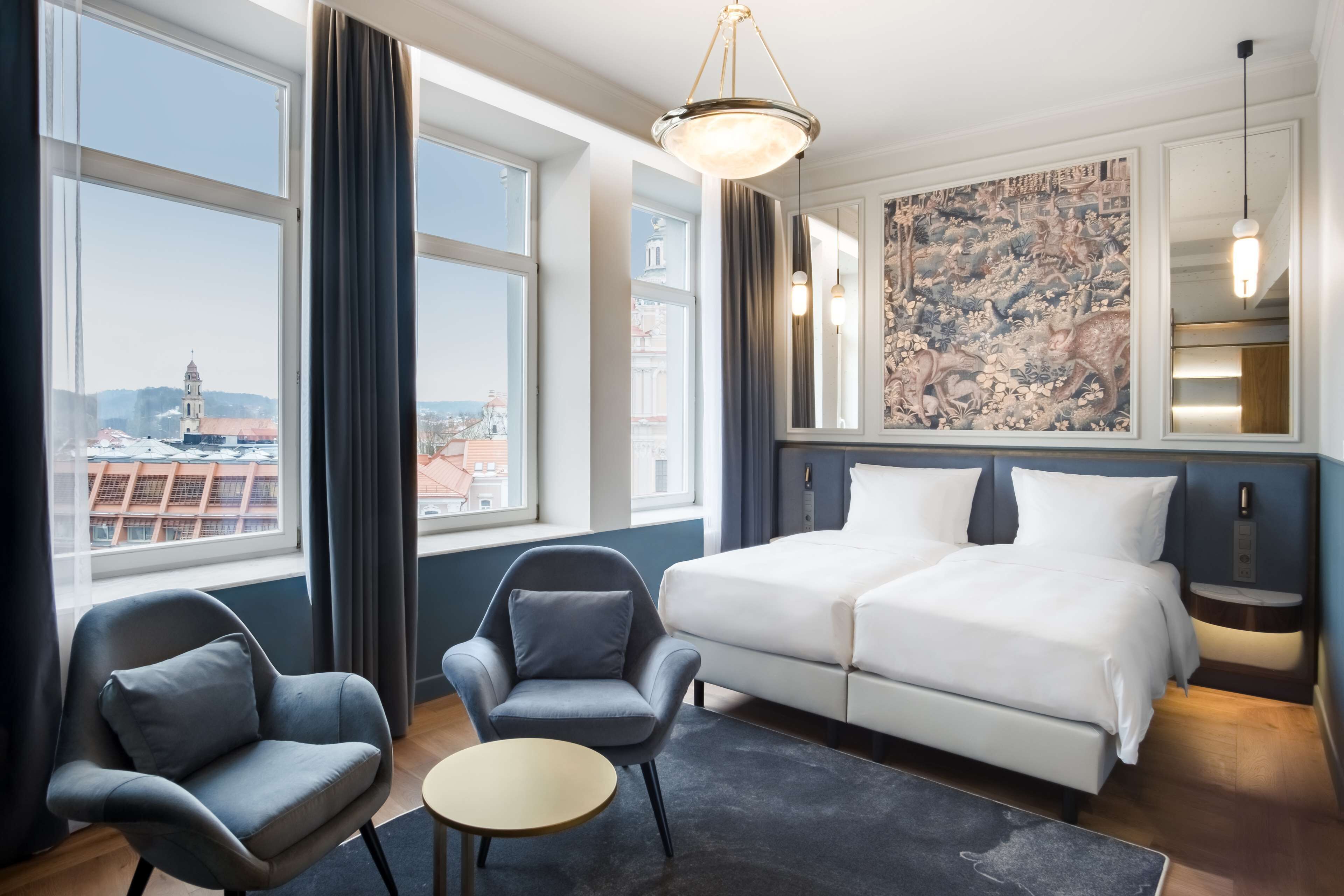 Een hotelkamer met een groot bed, twee fauteuils, een tafeltje en uitzicht op de stad door grote ramen.