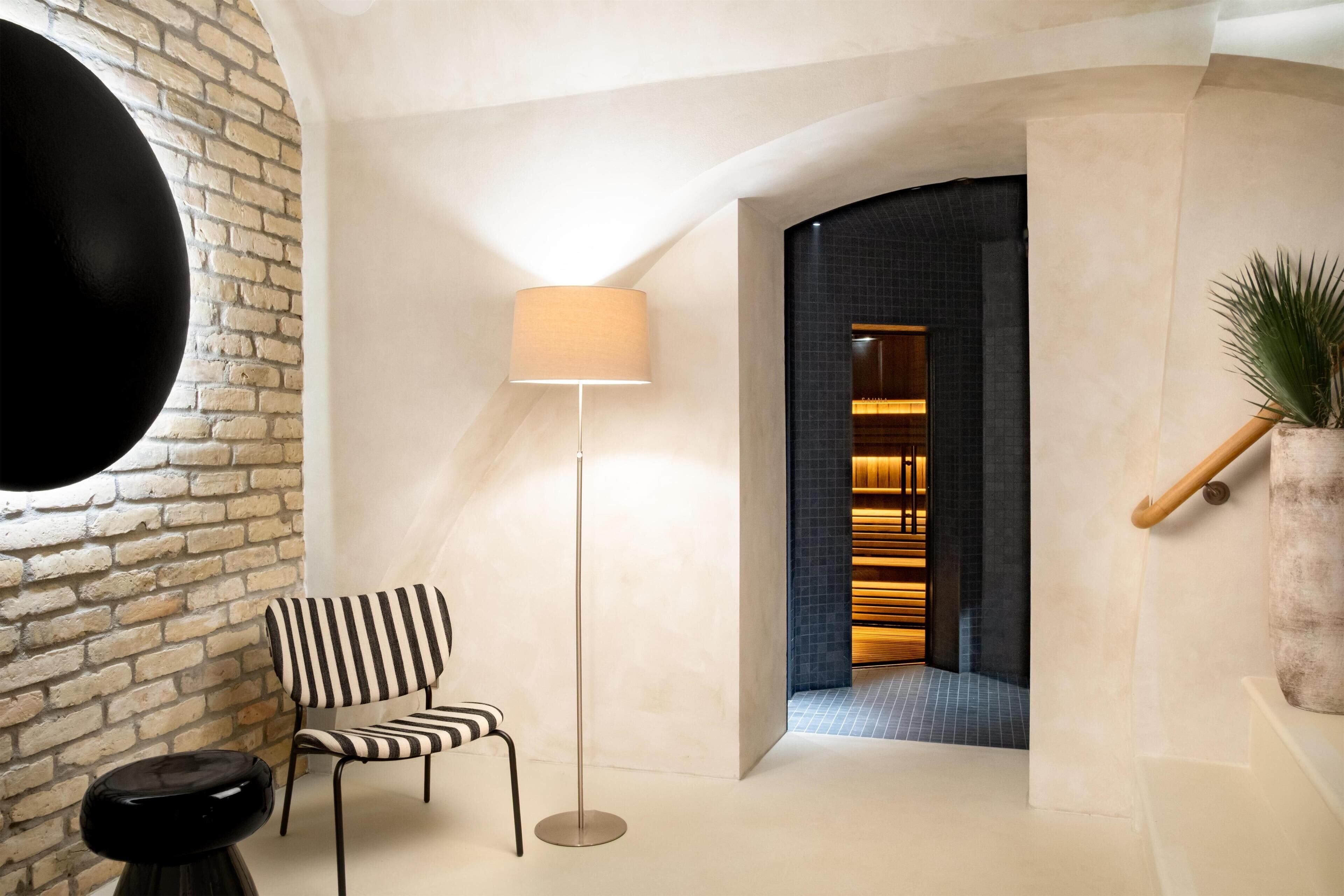 Ein modernes Interieur mit einer Ziegelwand, einem gestreiften Sessel, einer Stehlampe und einer Treppe, die zu einem dunkel gefliesten Bereich führt.