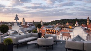 A rooftop terrace with outdoor seating, a decorative tree, and a view of the cityscape including historic buildings and a church. - Radisson Collection Astorija Hotel, Vilnius (Vilnius)