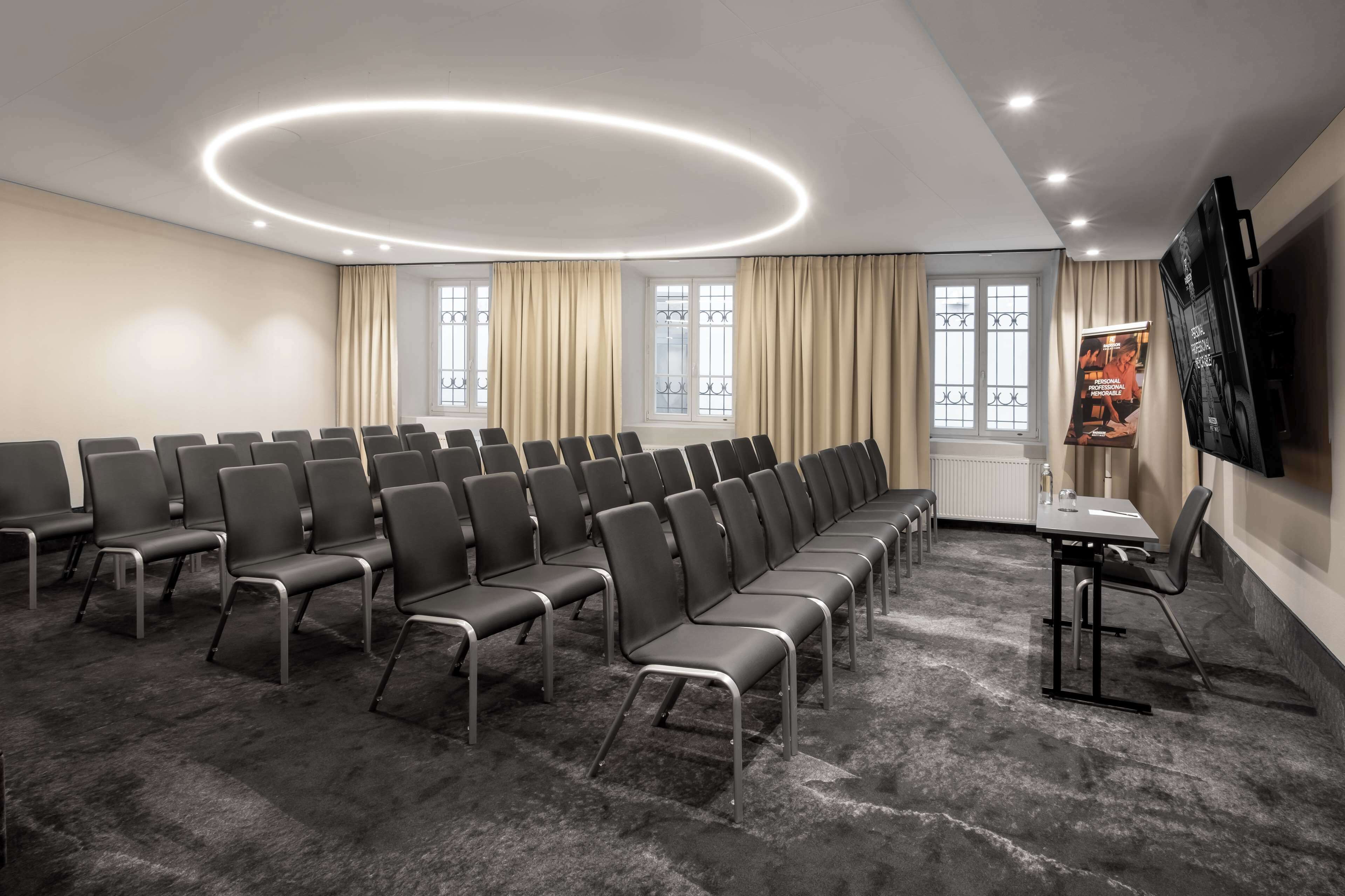 Un salón de conferencias con filas de sillas, una mesa, un televisor y ventanas con cortinas.