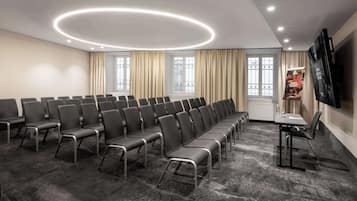 Un salón de conferencias con filas de sillas, una mesa, un televisor y ventanas con cortinas.