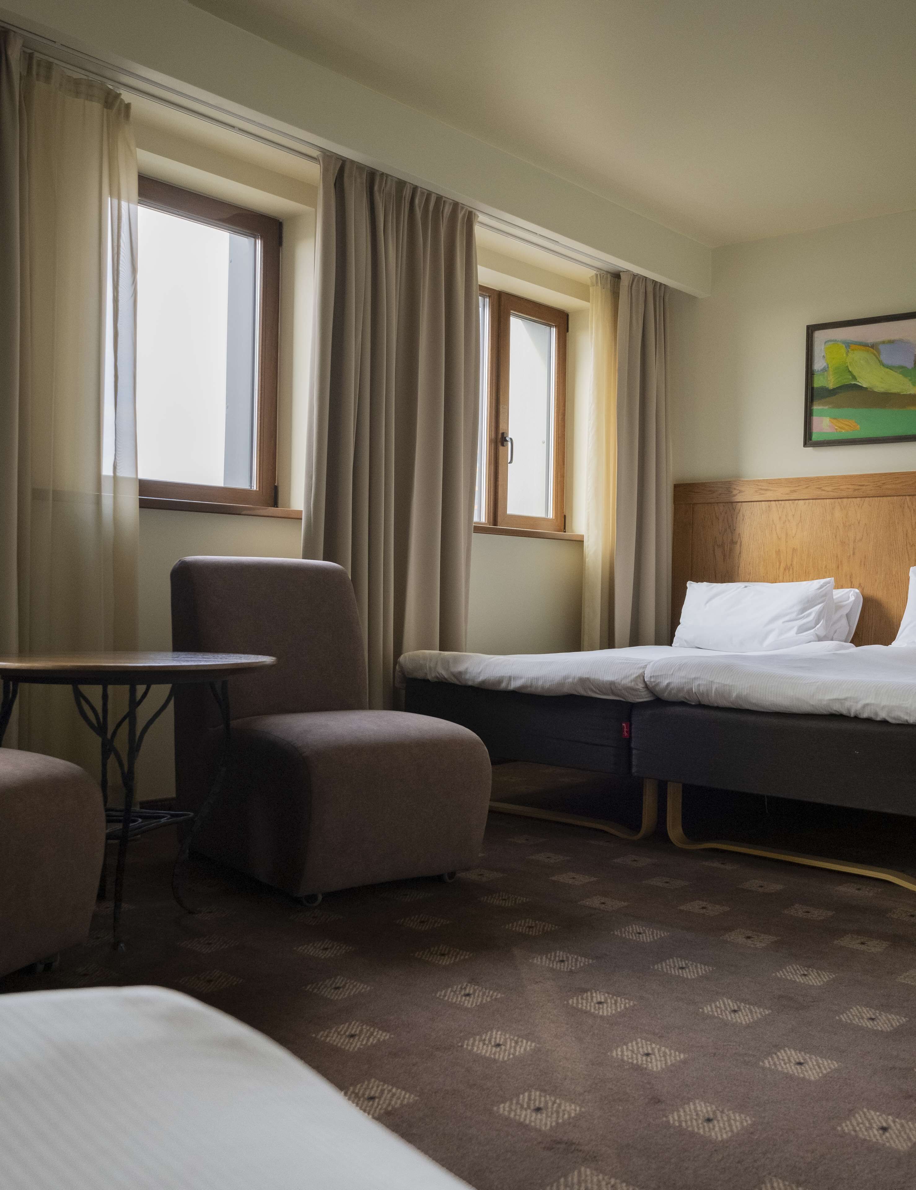 A hotel room with a bed, a chair, a small table, and two windows with curtains.