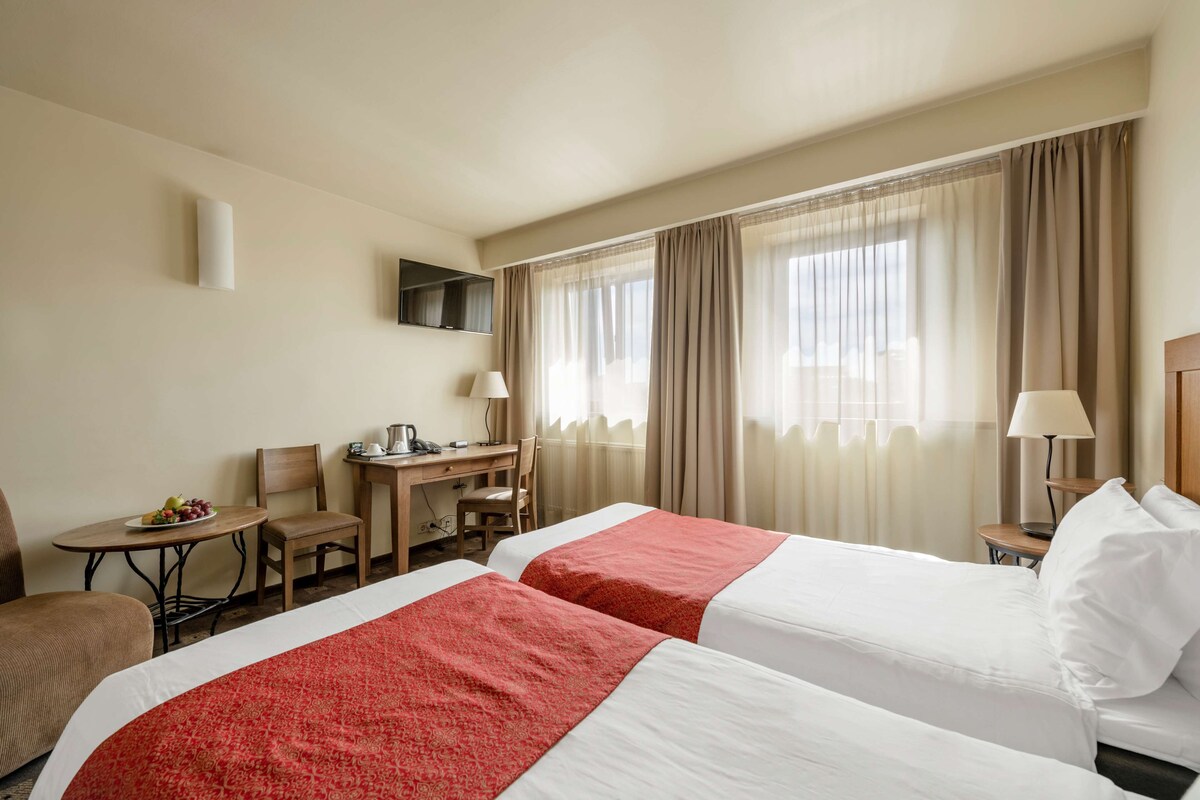 A hotel room with two beds, a desk, a chair, a small table with a fruit bowl, and a TV mounted on the wall.