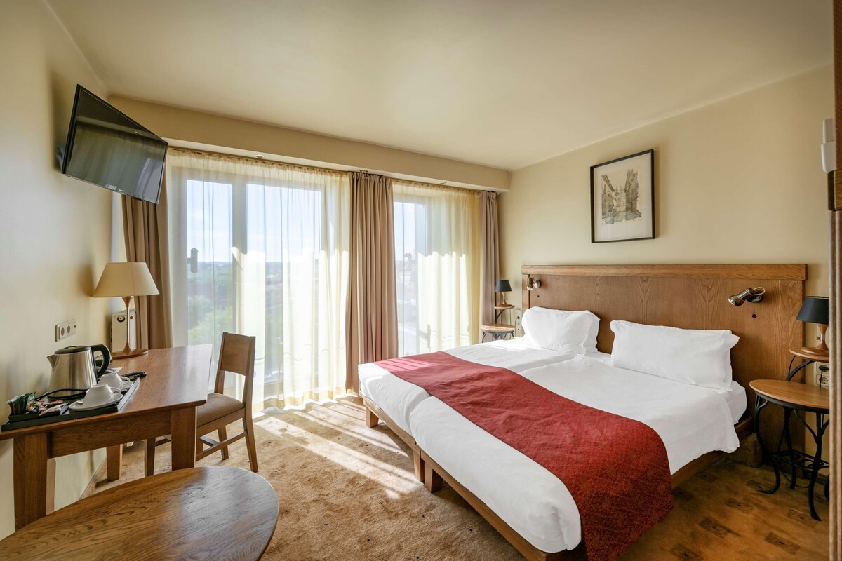Habitación de hotel con una cama grande, un escritorio con silla, un televisor montado en la pared y una ventana con cortinas.