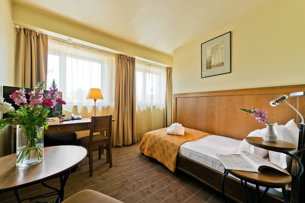 A hotel room with a bed, desk, chair, and a small table with a vase of flowers.