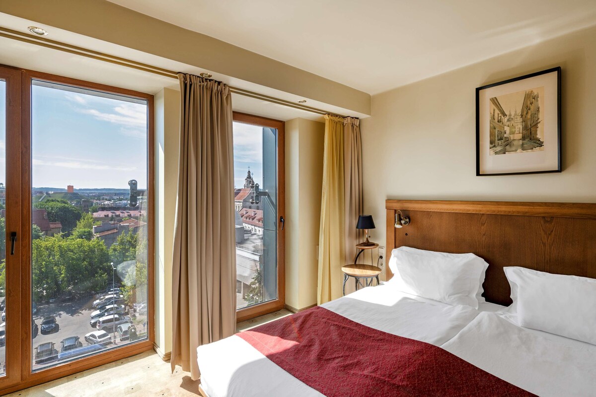 Un cuarto de hotel con una cama grande, vista de la ciudad a través de un ventanal corredero y un cuadro enmarcado en la pared.