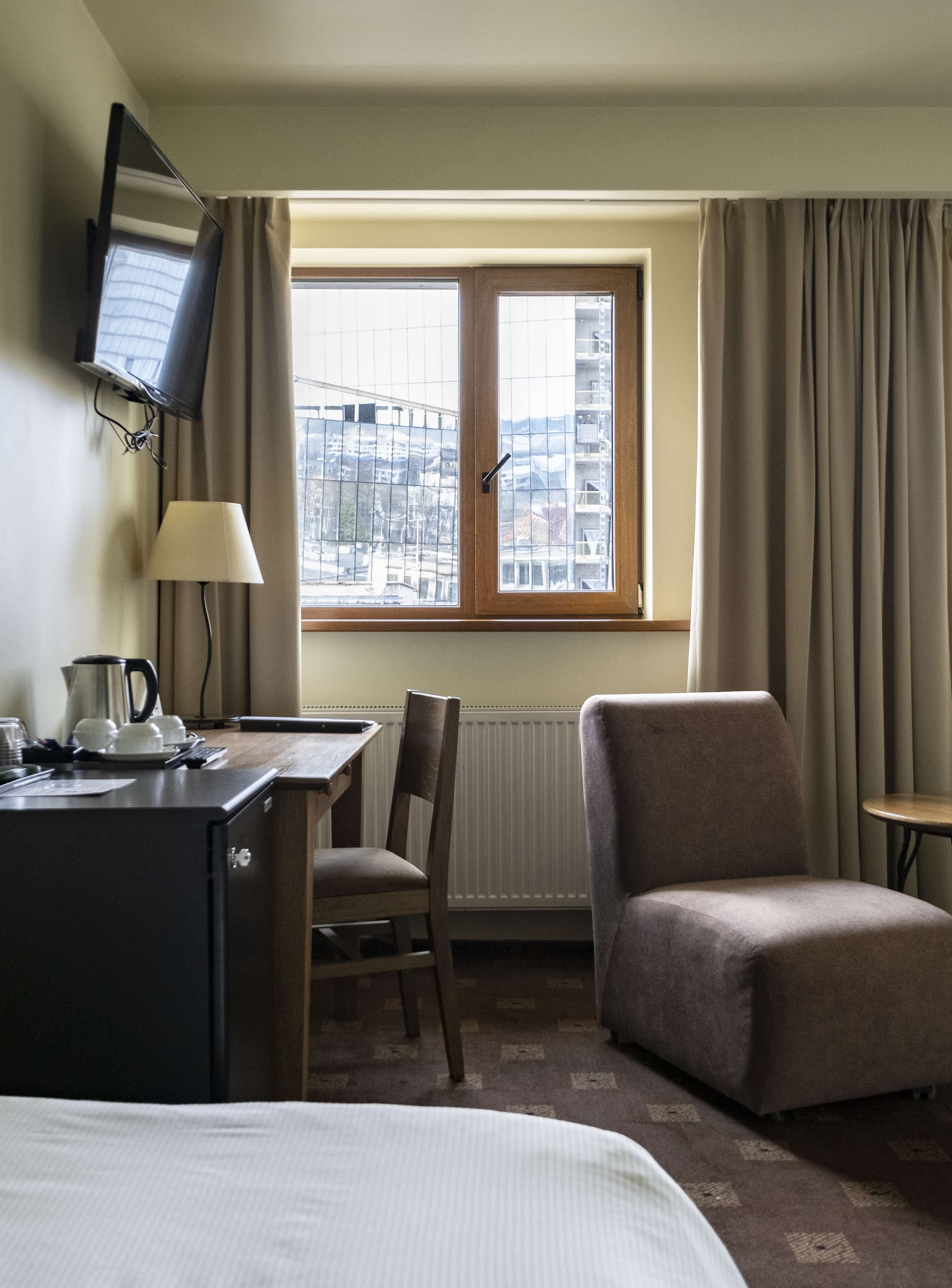 A hotel room with a bed, a desk with a lamp, a chair, a TV mounted on the wall, and a window with curtains.