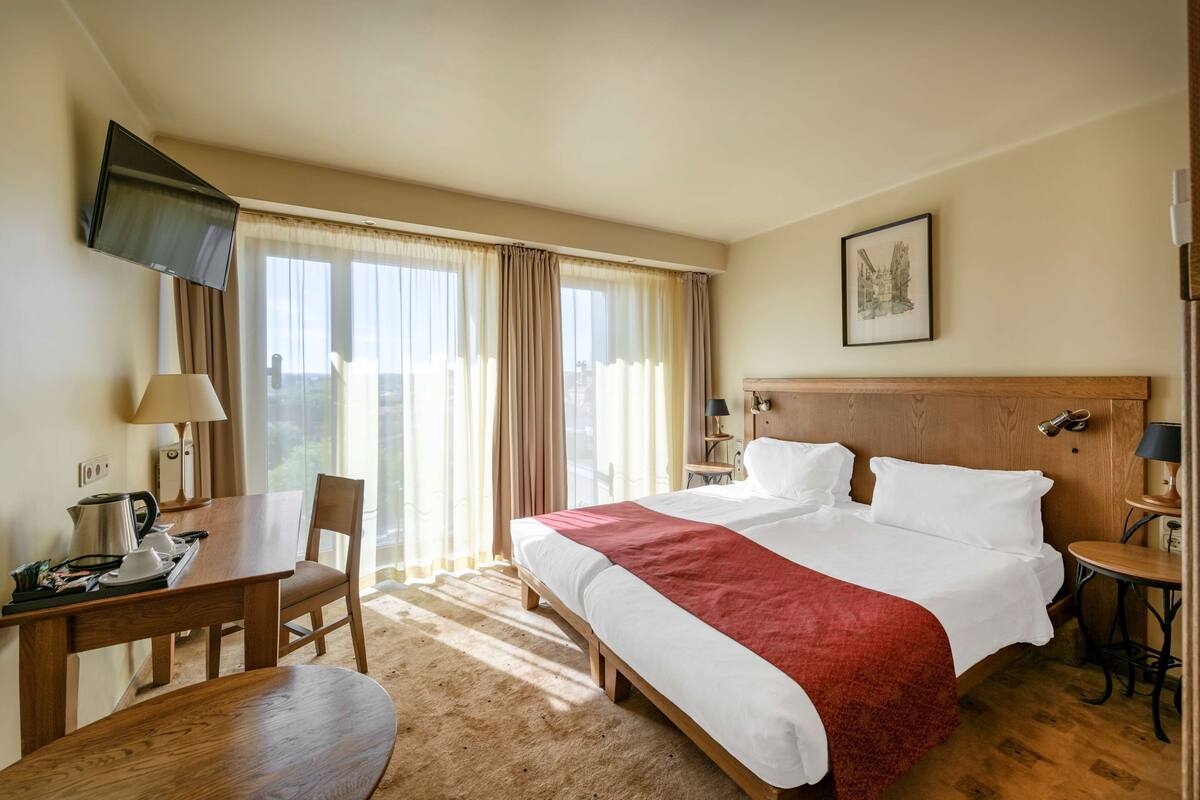 A hotel room with a bed, a desk, a chair, a TV mounted on the wall, and a window with curtains.