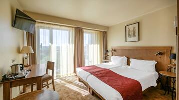 A hotel room with a bed, a desk, a chair, a TV mounted on the wall, and a window with curtains.