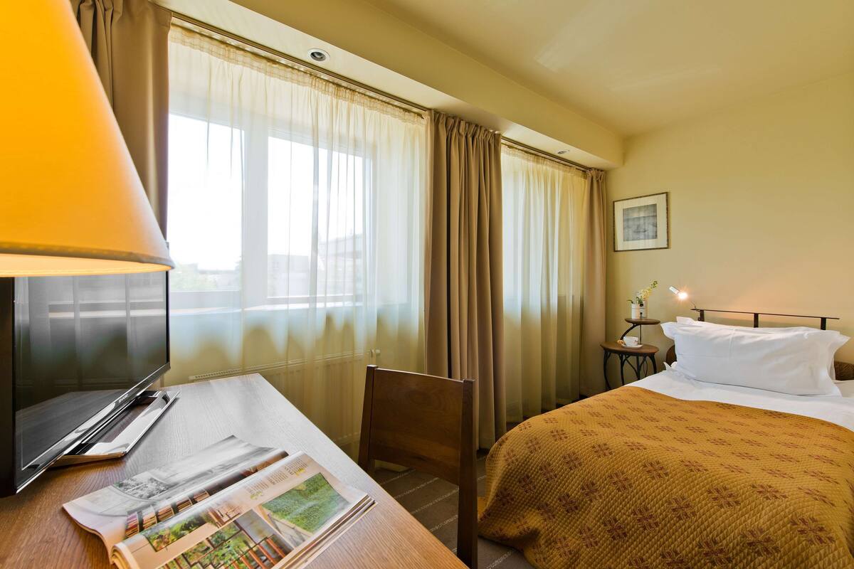 A hotel room with a bed, a desk with a lamp, a chair, a window with curtains, and a small table with a vase.