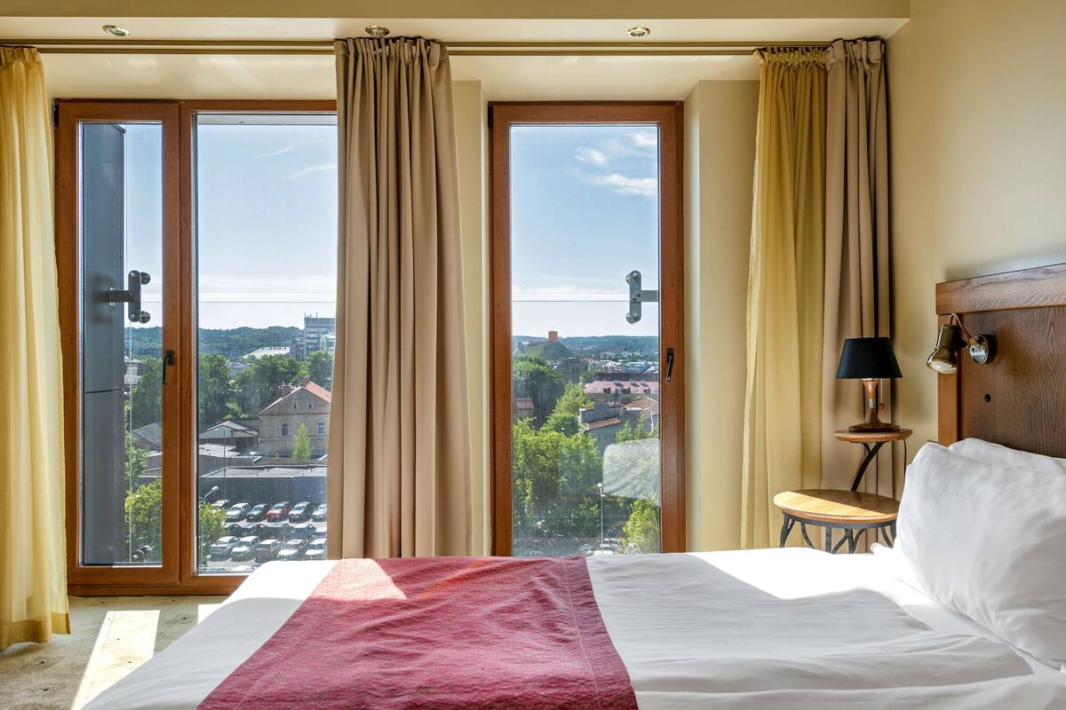 A hotel room with a large bed, a wooden headboard, a small table with a lamp, and a view of a cityscape through floor-to-ceiling windows.