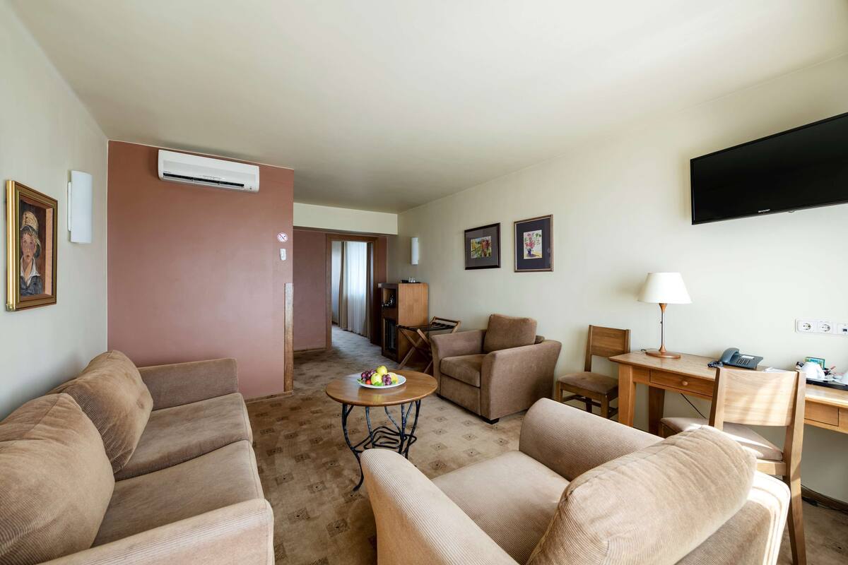 A living room with beige sofas, a small round table with fruit, a TV, and a desk with a lamp.