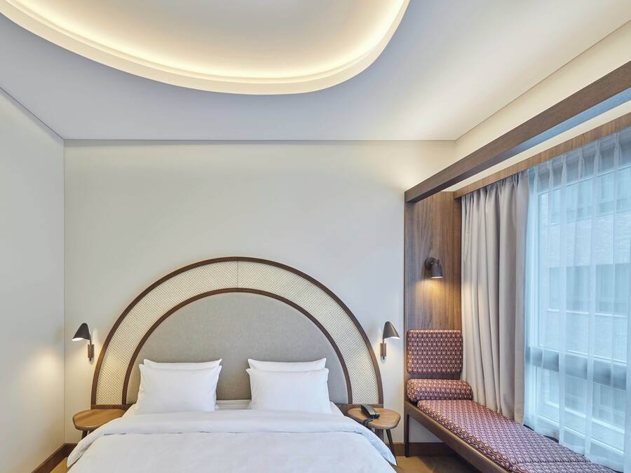 Moderni hotellihuone, jossa on suuri sänky, yöpöytä, pieni pöytä ja ikkuna, jossa on verhot.
