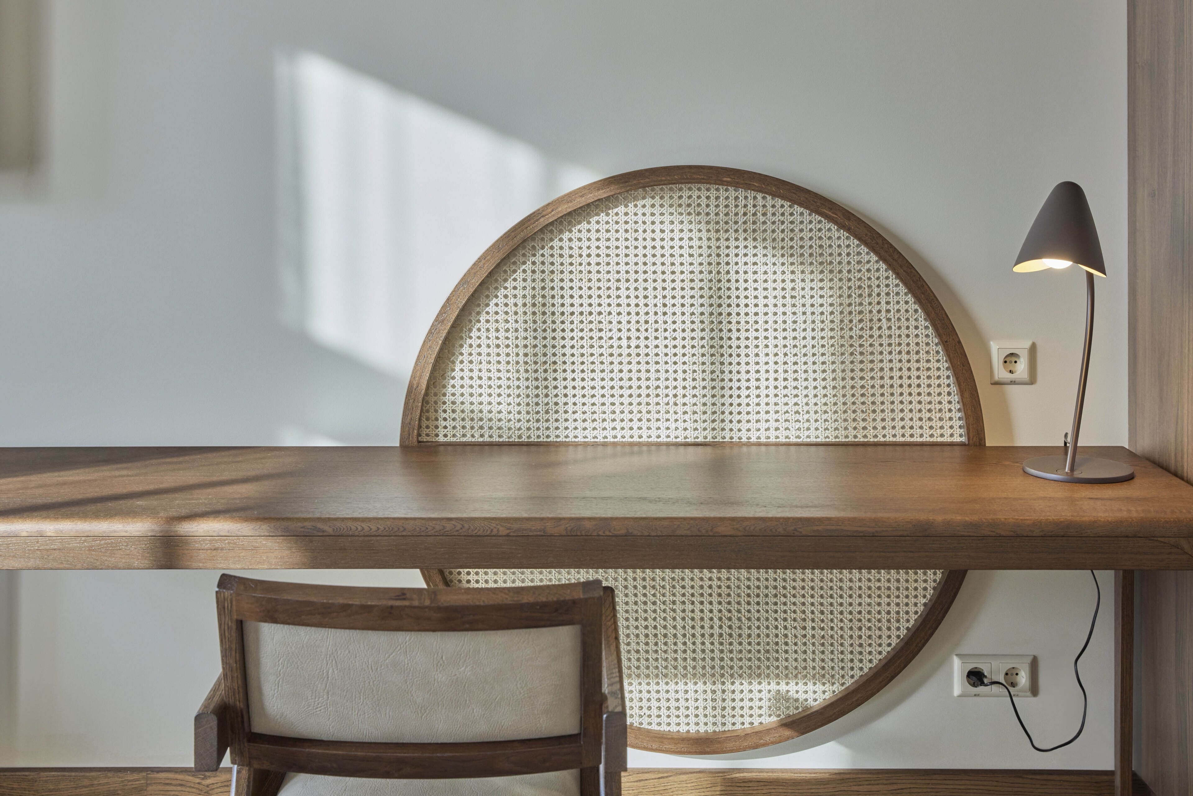 A wooden desk with a round woven backrest, a chair, and a desk lamp.