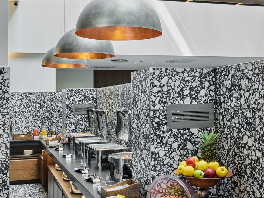 A modern kitchen with marble walls, a long counter with various food items, and pendant lighting.