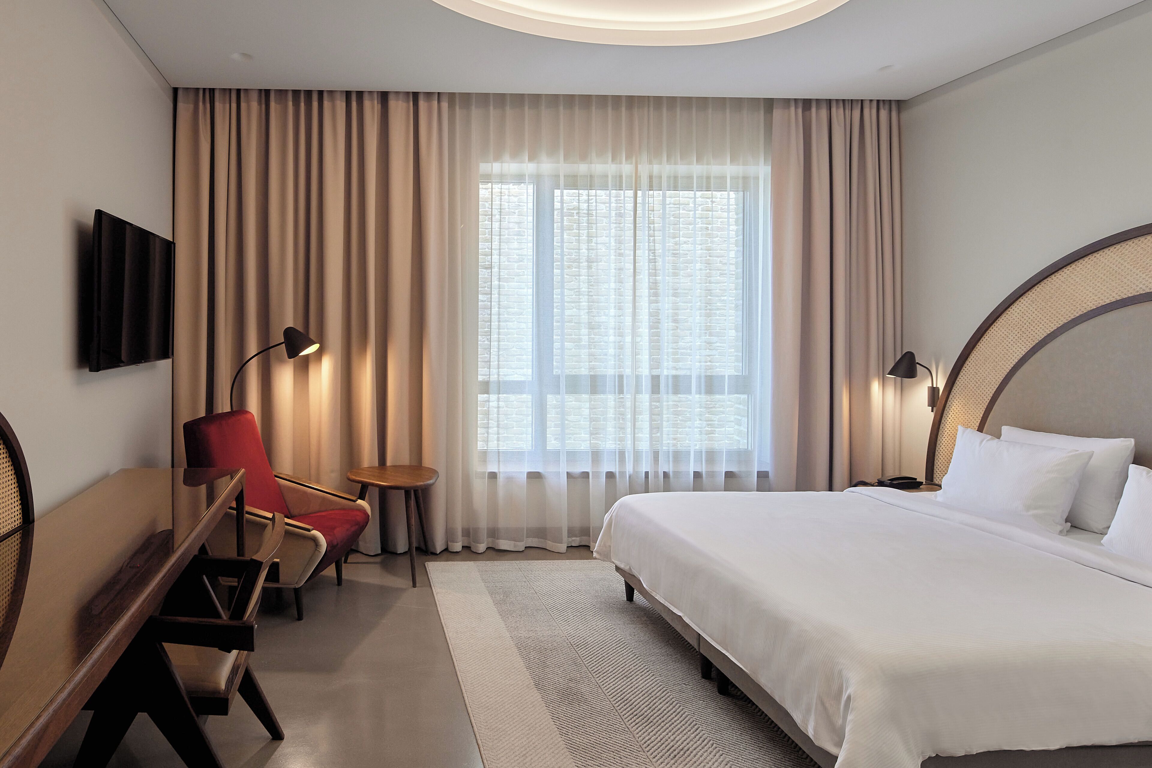 A hotel room with a large bed, a desk with a chair, a TV mounted on the wall, and a window with sheer curtains.