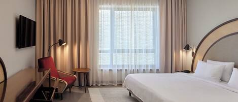 A hotel room with a large bed, a desk with a chair, a TV mounted on the wall, and a window with sheer curtains.