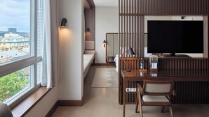 A modern hotel room with a large window, a bed, a desk with a chair, a TV, and a view of the cityscape. - Neringa Hotel (Vilnius)