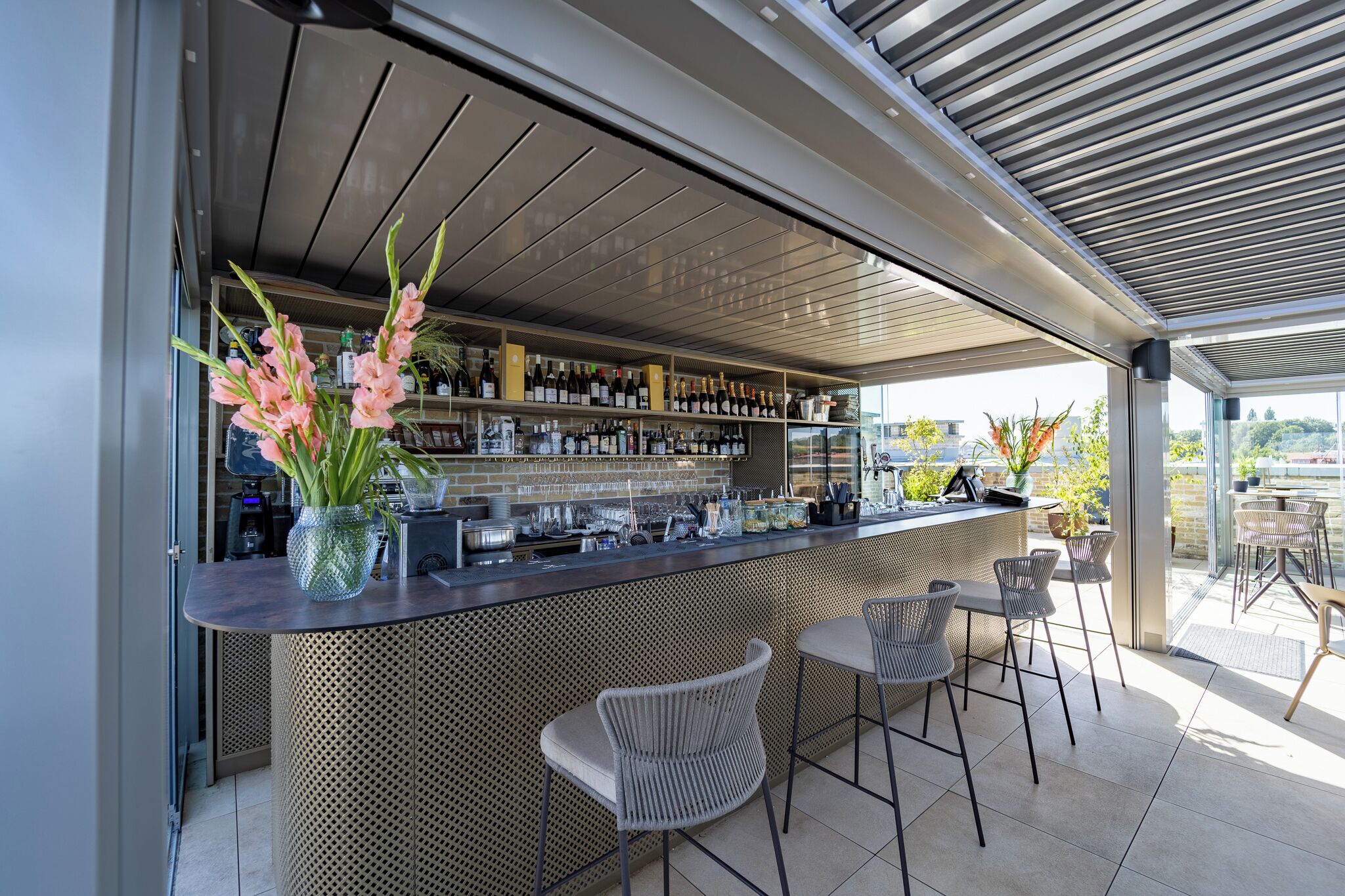 Un'area bar esterna moderna con tetto in metallo, sgabelli da bar e diverse bottiglie e bicchieri sugli scaffali.