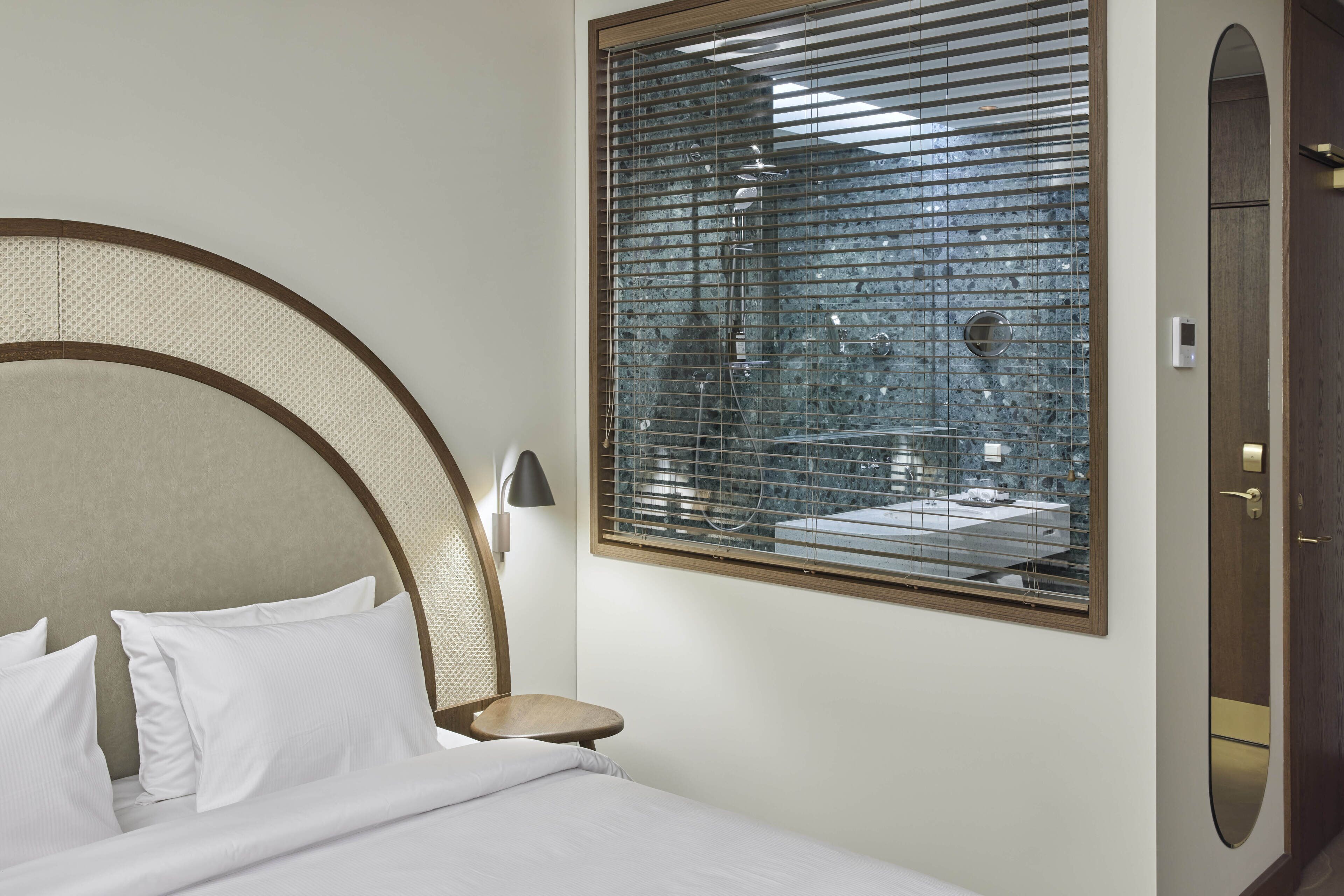 Quarto de hotel com uma cama grande, uma mesinha, luminária na parede e janela com vista para um banheiro.