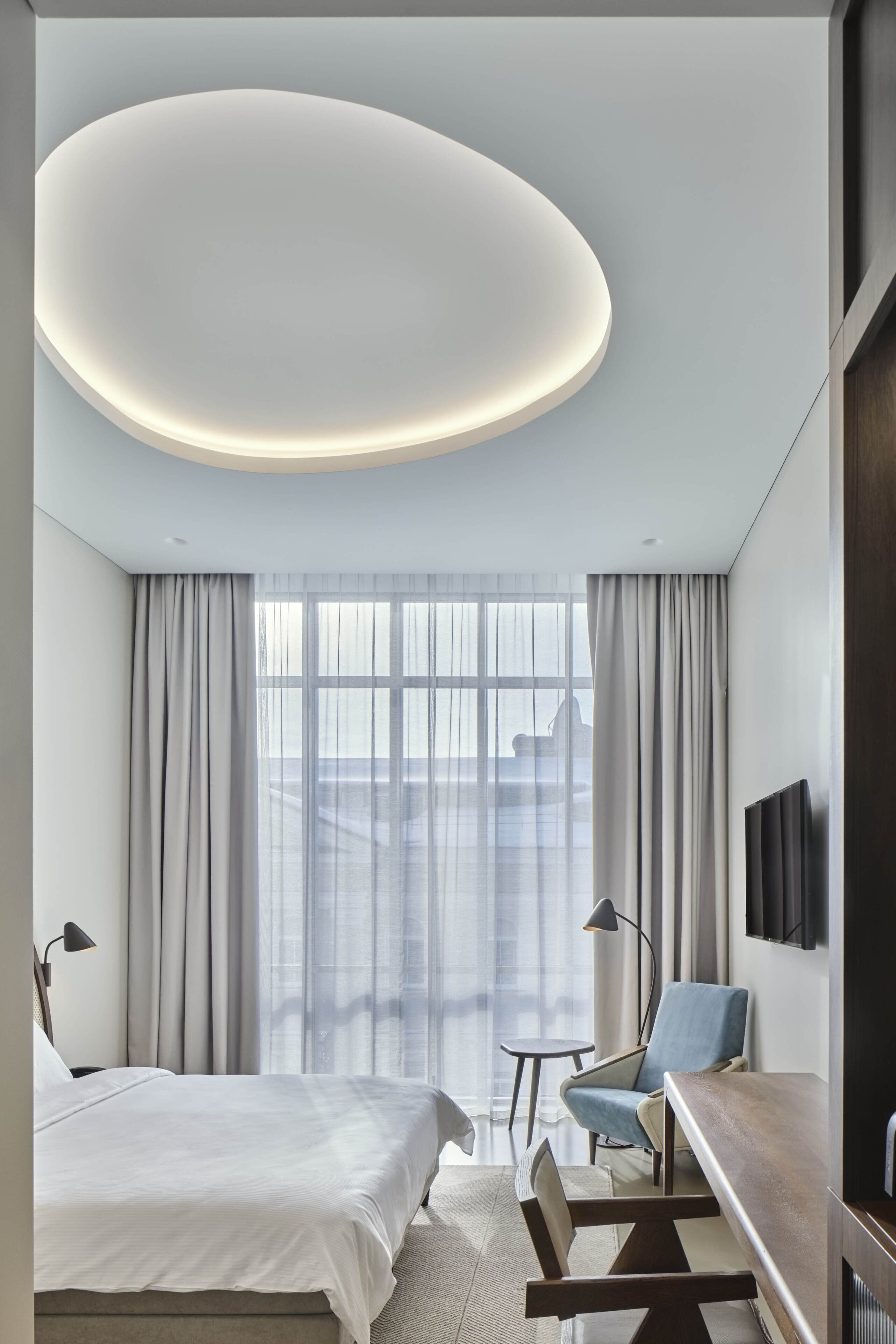 A modern hotel room with a large bed, a desk, a chair, a TV, and a large window with curtains.