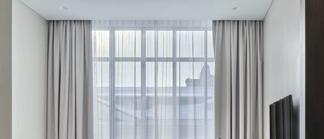 A modern hotel room with a large bed, a desk, a chair, a TV, and a large window with curtains.