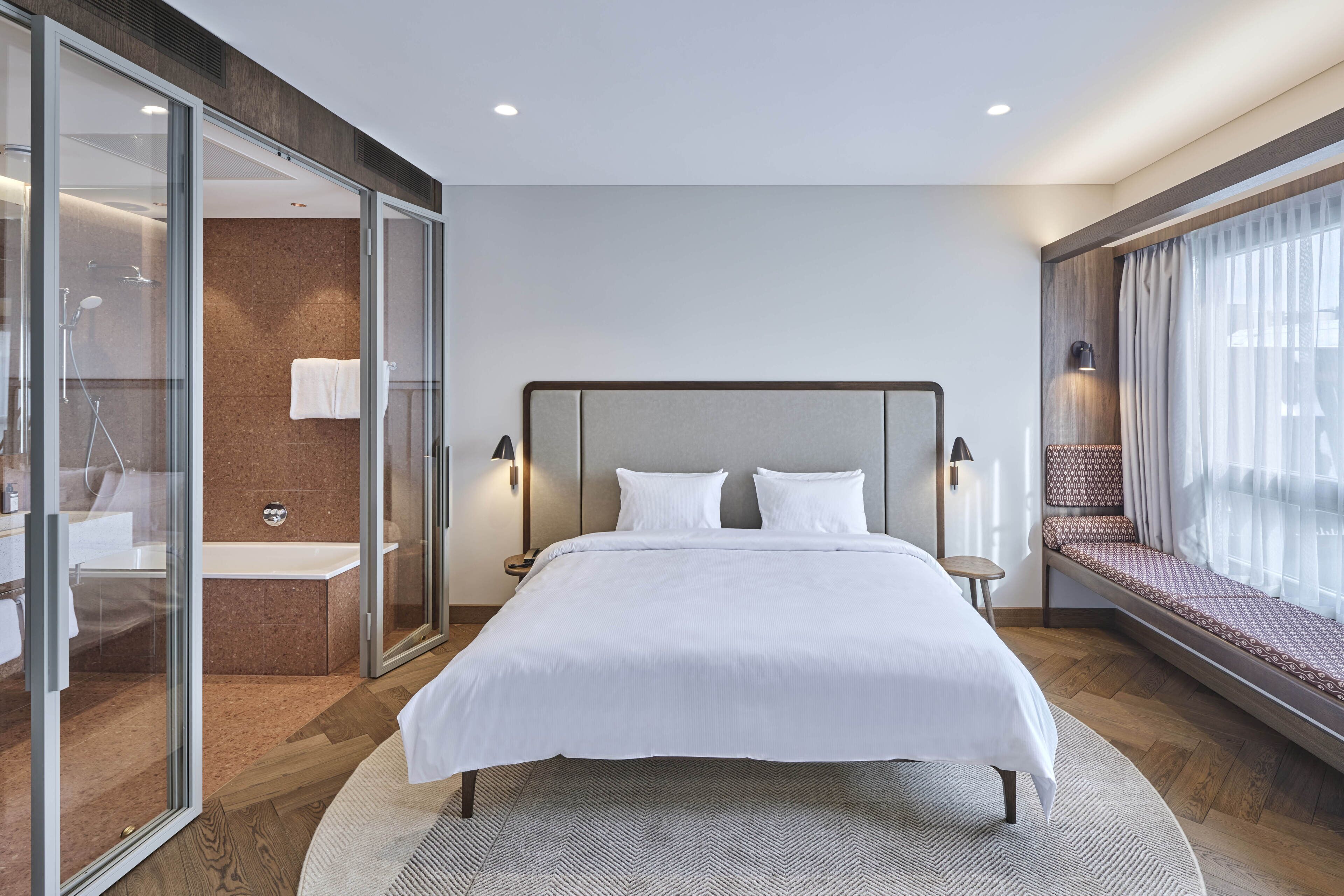 A modern hotel room with a large bed, a glass shower enclosure, a built-in bench with a cushioned seat, and a view of the city through large windows.
