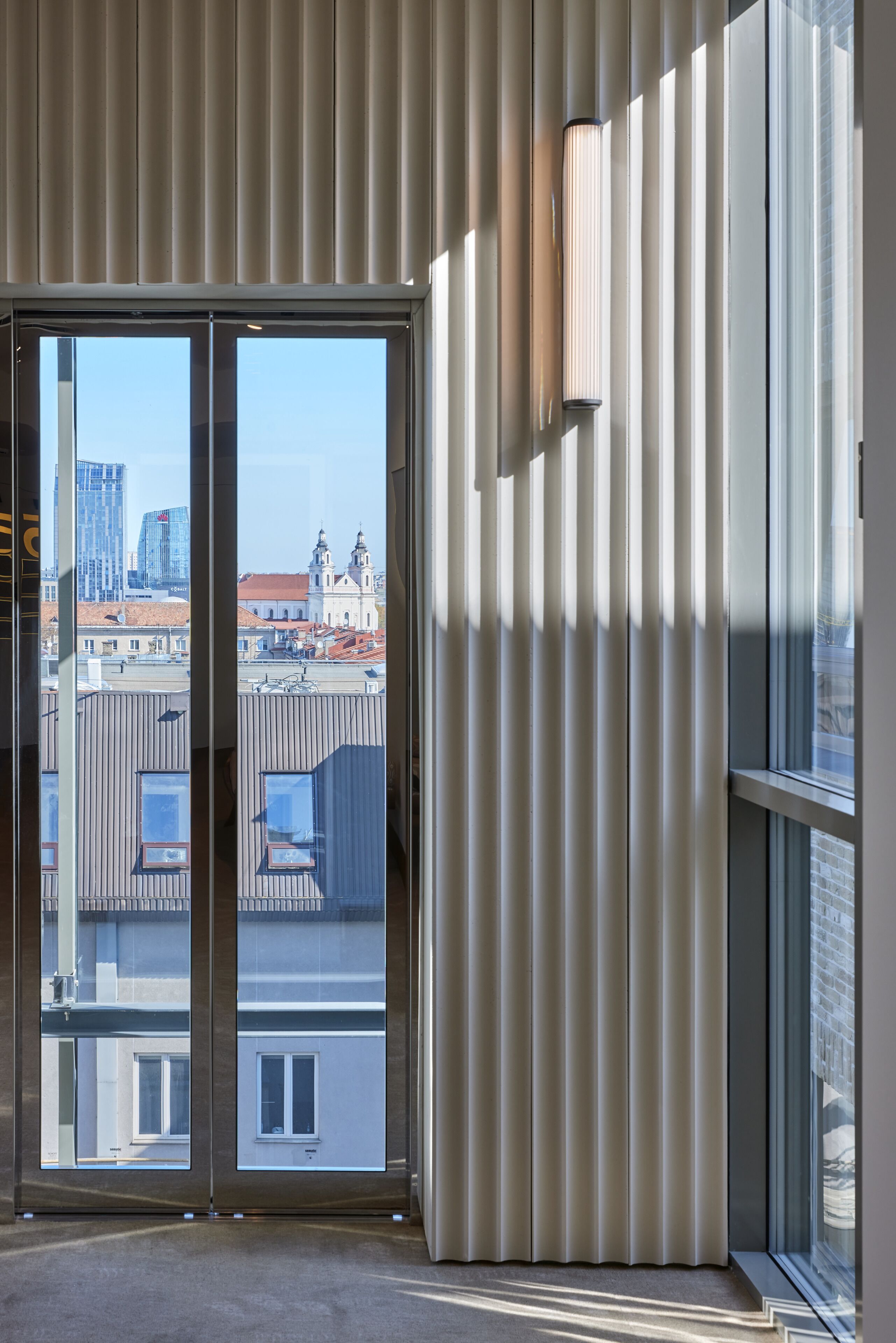 A modern interior with large windows, a view of buildings, and a vertical light fixture.
