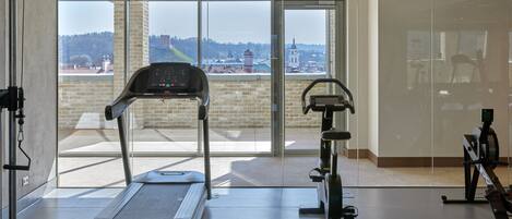 A gym with a treadmill, stationary bike, and rowing machine. It has large windows with a view of buildings and trees.