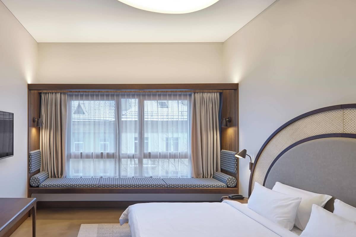 A hotel room with a large bed, a window seat with curtains, a round headboard, and a bedside table.