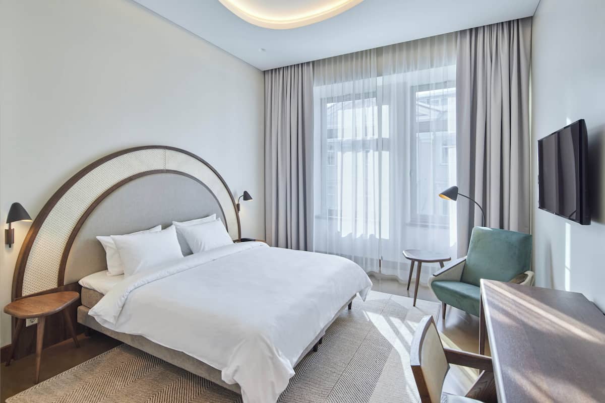 A modern hotel room with a large bed, a desk, a chair, a TV, and a window with curtains.