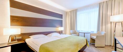 A hotel room with a large bed, two bedside tables with lamps, a small round table, two chairs, and a large window with curtains.