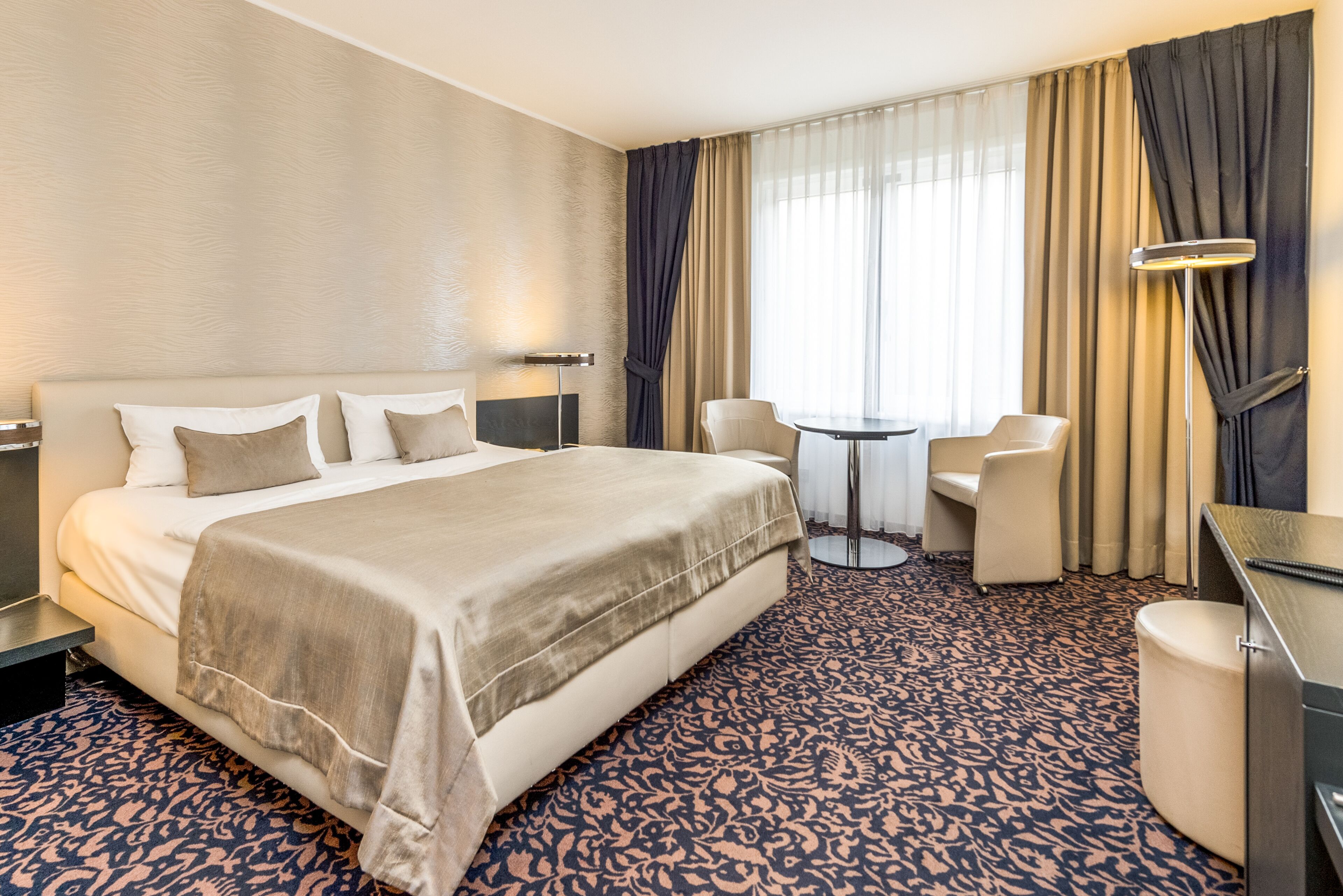 A hotel room with a large bed, a desk, a chair, a small table, and a lamp.