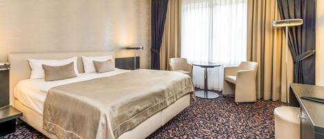 A hotel room with a large bed, a desk, a chair, a small table, and a lamp.