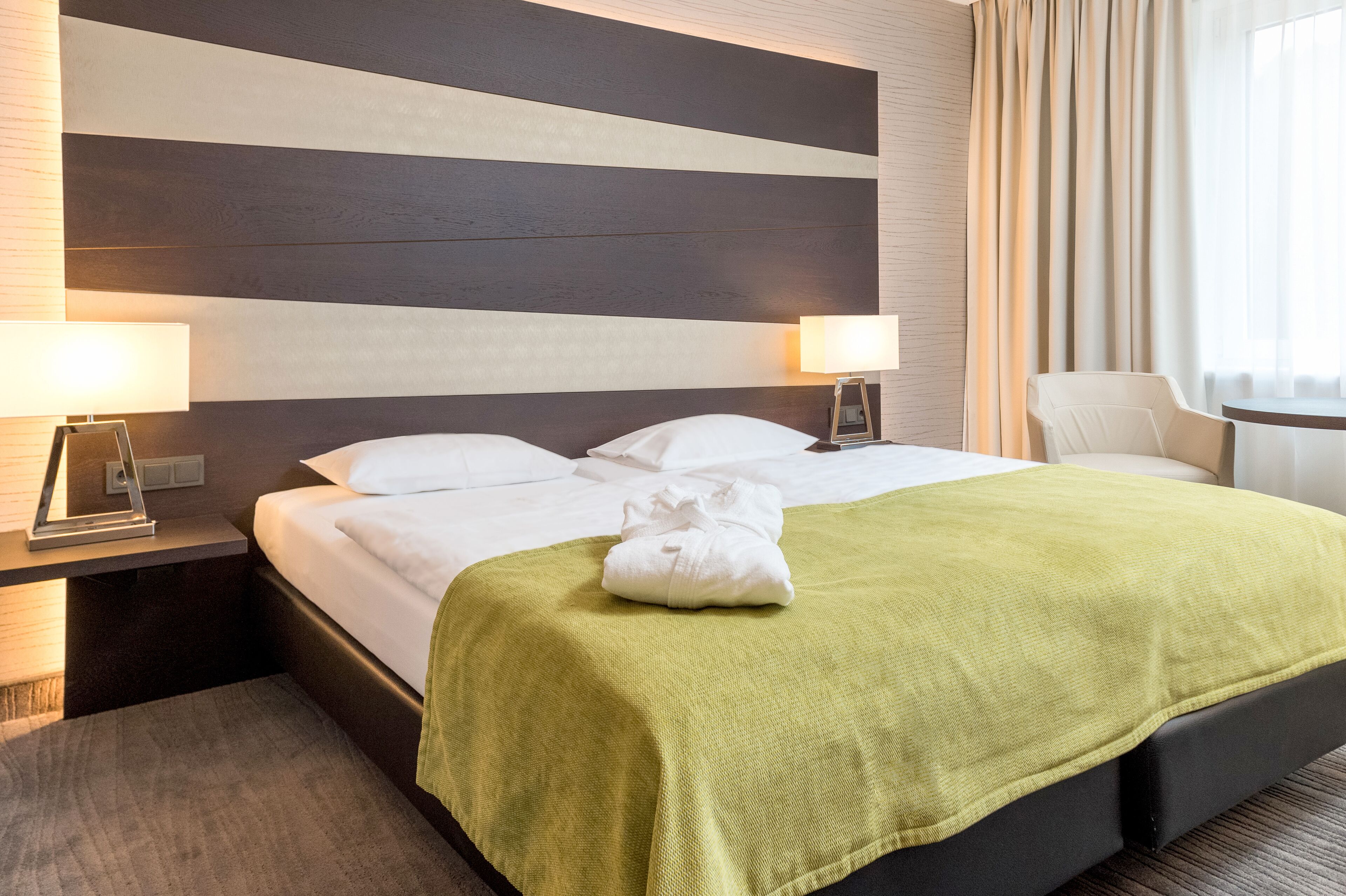 Une chambre d’hôtel avec un grand lit, deux lampes de chevet, un plaid vert et une vue sur l’extérieur par une fenêtre avec des rideaux.