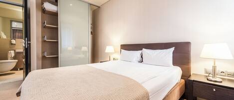 A modern hotel room with a large bed, a nightstand with a lamp, a bathroom with a bathtub, and a sliding closet door.