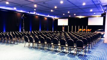 Un salón de conferencias con filas de sillas, un escenario con un atril y dos pantallas de proyección grandes.