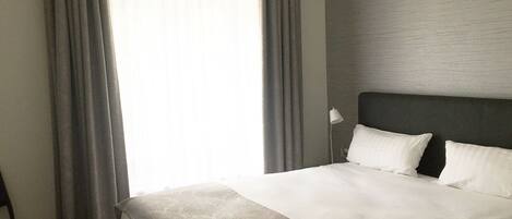 Habitación de hotel con una cama grande, cortinas grises, una silla y una lámpara.
