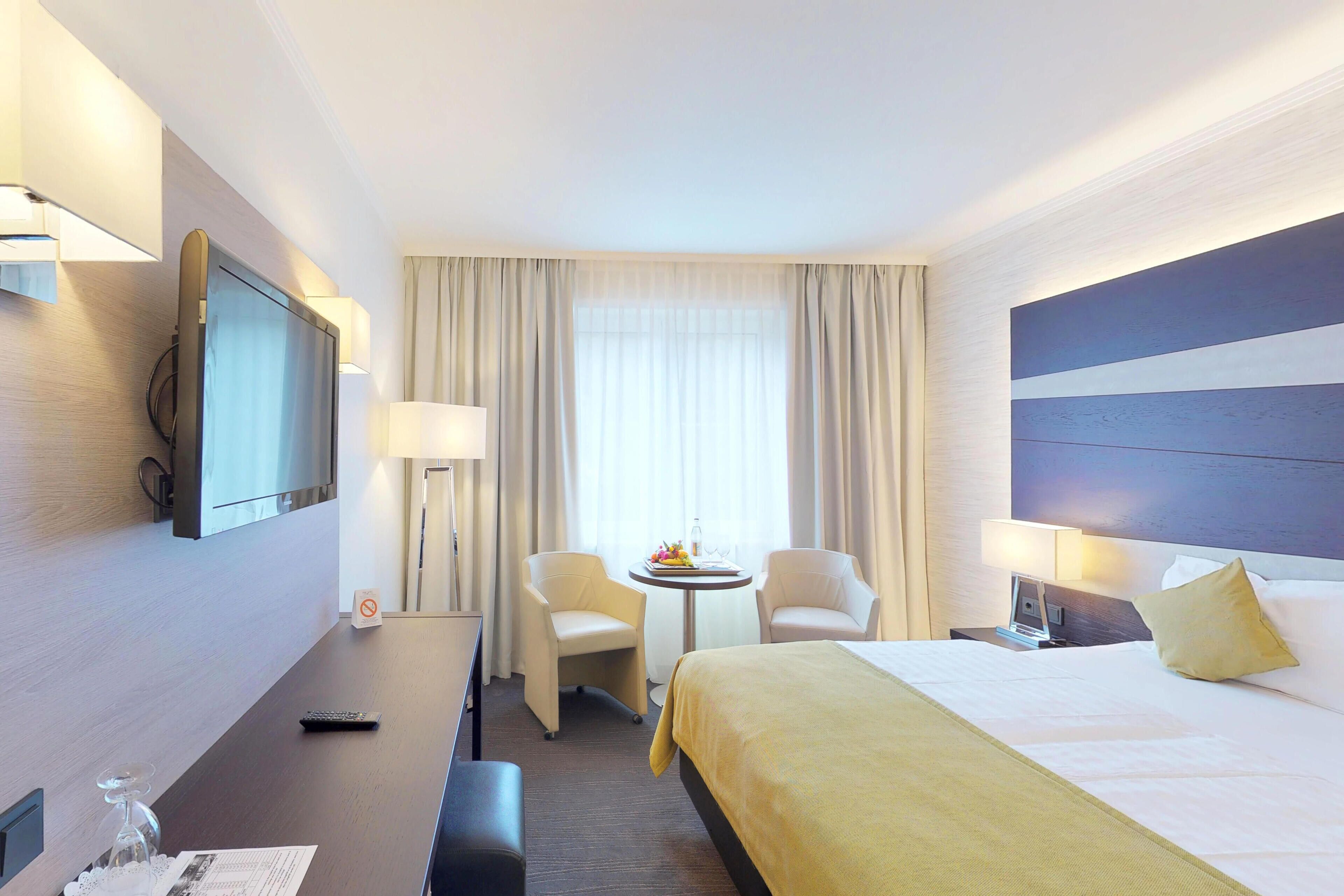 Habitación de hotel con cama, dos sillas, una mesita con lámpara, televisión y ventana con cortinas.