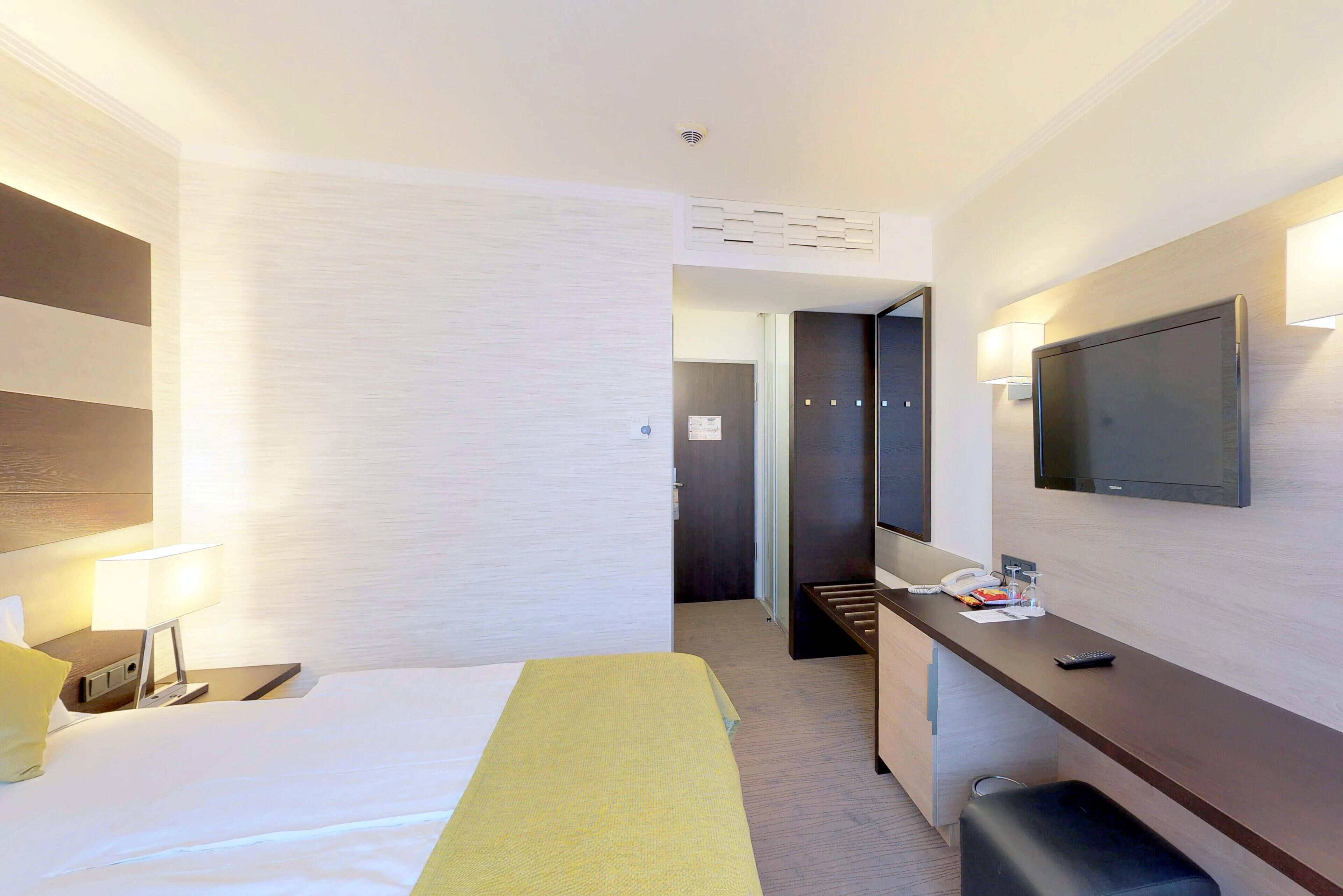 Een hotelkamer met een bed, nachtkastje, lamp, televisie en een bureau met stoel.