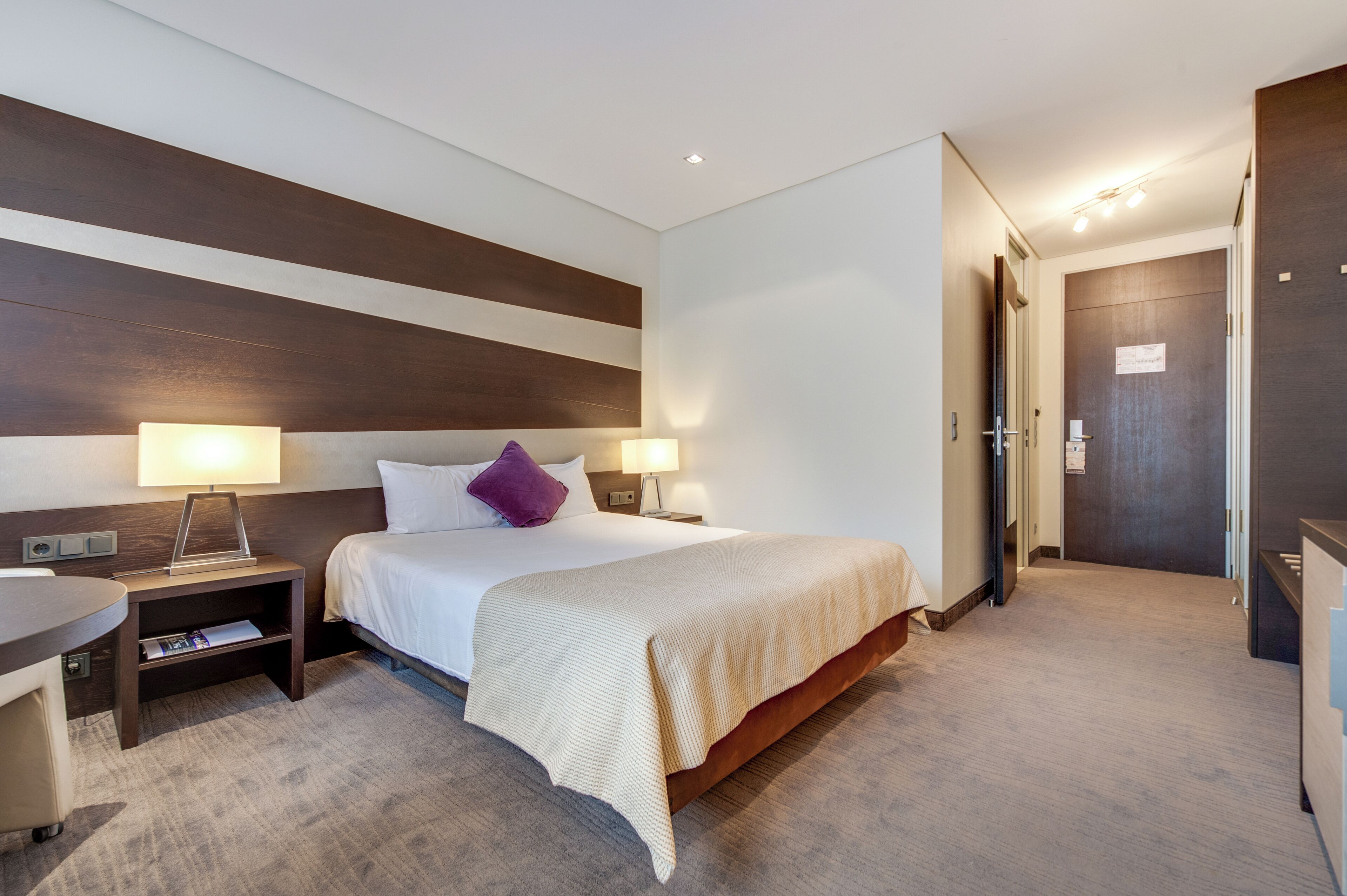 Une chambre d’hôtel avec un grand lit, deux tables de chevet avec des lampes, un petit bureau et une porte donnant sur une autre pièce.