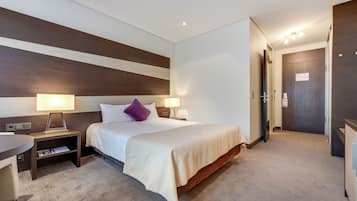 A hotel room with a large bed, two bedside tables with lamps, a small desk, and a door leading to another room.
