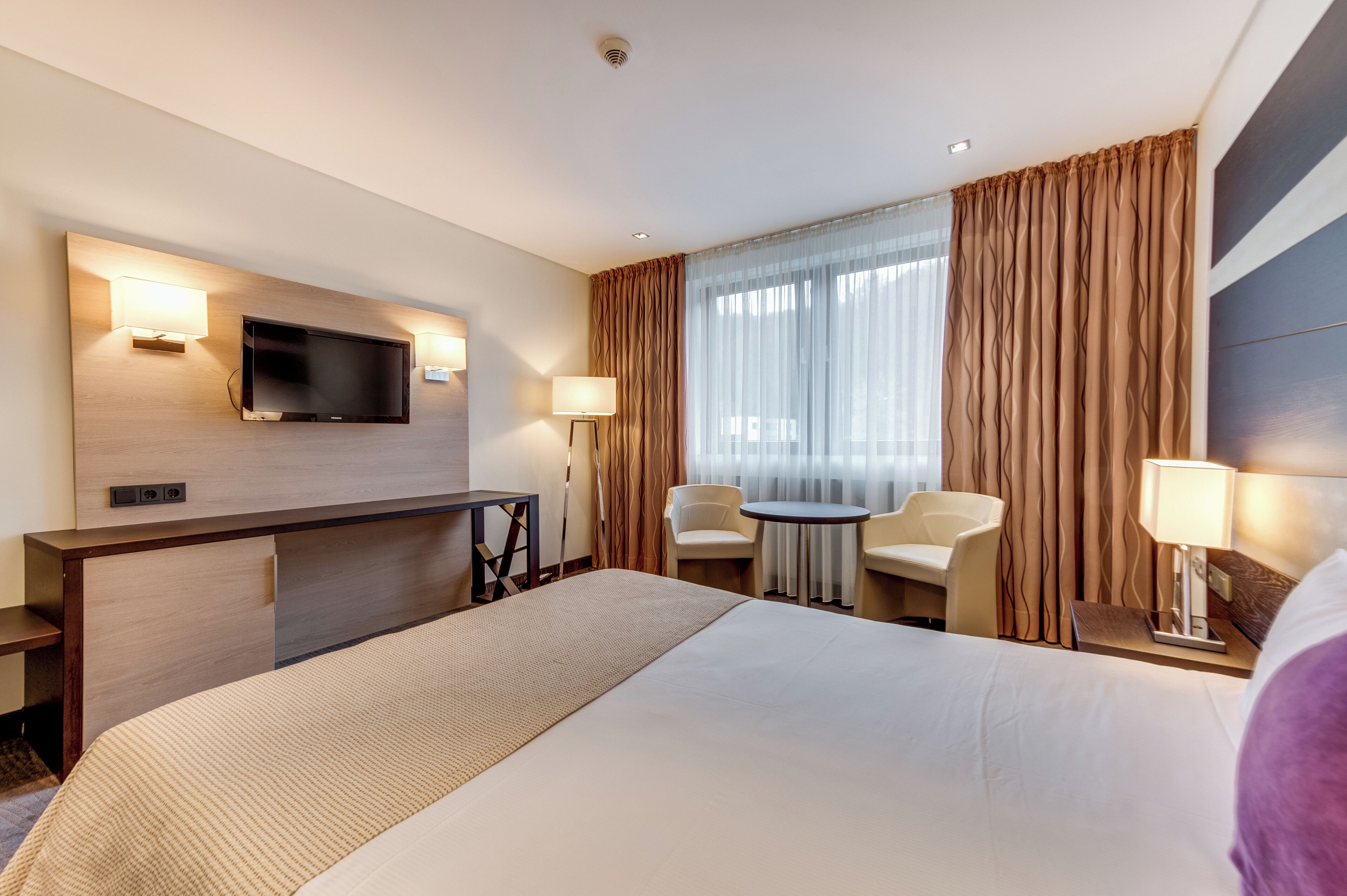 Una habitación de hotel con cama, escritorio, televisor, dos sillas y ventana con cortinas.