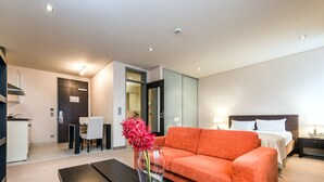 A modern hotel room with a sofa, a dining table, a kitchen area, and a bed with a nightstand. - Parc Hotel Alvisse (Luxembourg City)
