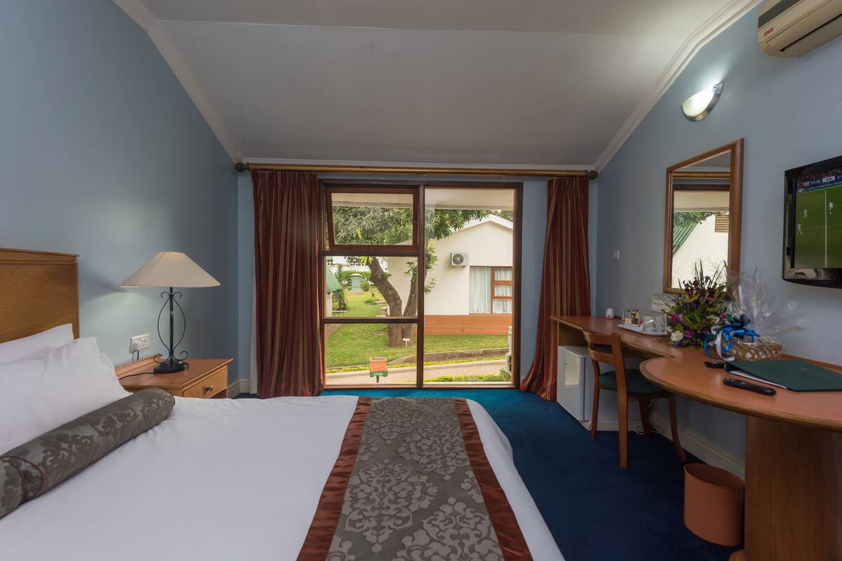 A hotel room with a bed, a desk, a chair, a lamp, and a view of the outside.